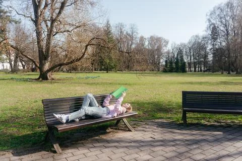 Student relaxing on a bench while reading a book outdoors on a sunny day 스톡 사진
