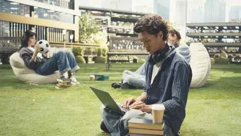 Student smiling and writing notes on laptop while relaxing on grass Stock Photos