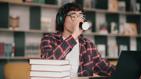 Student studying with laptop and coffee in library wearing headphones and Stock Footage 341889329