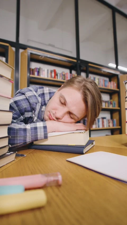 Student studying in a library with books piled up and falling asleep on the Stock Footage 318089643