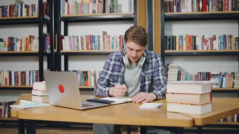 Student studying at a table in a library while taking notes and using a laptop Stock Footage 318089946