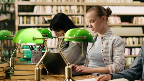 Student typing on laptop and taking notes from open book at library desk Stock Footage 336360476