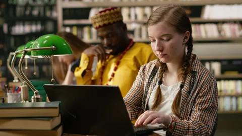 Student typing on laptop and taking notes beside open book at busy study table Stock Footage 337231881