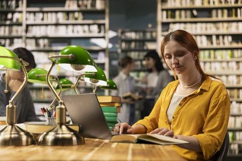 Student using laptop and open book at library desk with green study lamps Foto stock