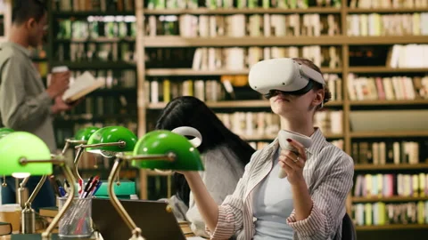 Student using virtual reality headset and hand controllers at library desk Stock Footage 336360570