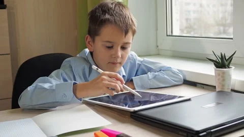 A student writes with a pencil on a tablet. a boy in a blue shirt is sitting at Stock Footage 169358814