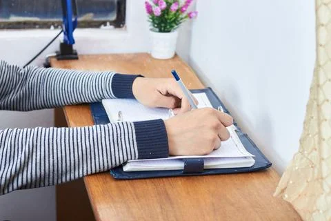 Student writing notes in spiral notebook with blue folder on wooden desk Stock Photos