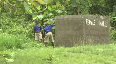 STUDENTS CUTTING GRASS WITH MACHETES BY OUTDOOR TOILETS MARKED MALE AND FEMALE Stock Footage 60116219