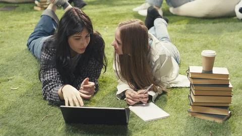 Students lay on grass beside textbook and laptop while discussing project ideas Stock Photos