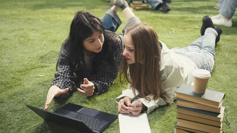 Students lay on grass beside textbook and laptop while discussing project ideas Stock Photos