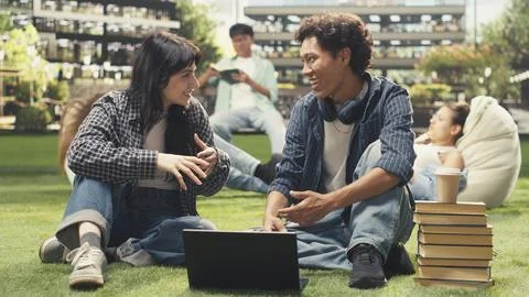 Students sitting face to face on grass with open textbook and stacked books Stock Photos