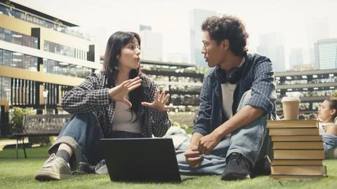 Students sitting face to face on grass with open textbook and stacked books Stock Photos