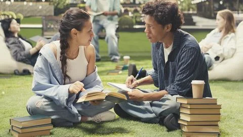 Students taking notes sitting on grass beside stacked books on campus Stock Photos