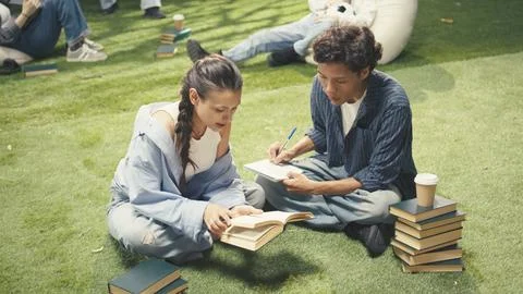 Students taking notes sitting on grass beside stacked books on campus Stock Photos