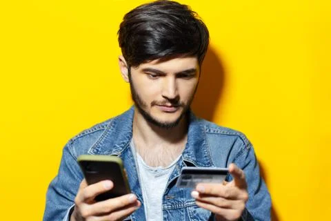 Studio portrait of young guy using smartphone and credit card, on yellow. Stock Photos