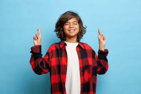 Studio shot of positive curly haired boy, keeps fingers crossed, makes a wish Stock Photos