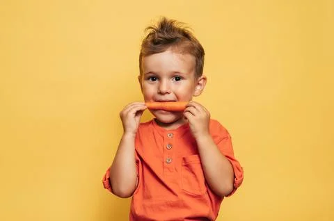 Studio shot of a smiling boy eating fresh carrots on a yellow background. The Stock Photos