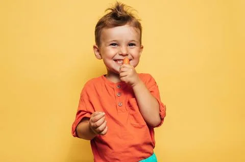 Studio shot of a smiling boy eating fresh carrots on a yellow background. The Stock Photos
