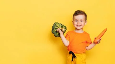 A studio shot of a smiling boy holding fresh broccoli and carrots on a yellow Stock Photos