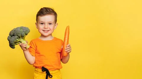 A studio shot of a smiling boy holding fresh broccoli and carrots on a yellow Stock Photos