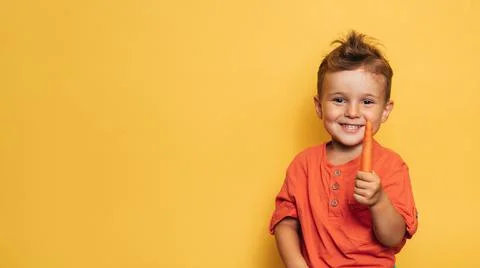 Studio shot of a smiling boy holding a fresh carrot on a yellow background. The Stock Photos
