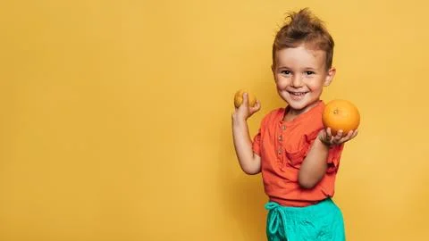 Studio shot of a smiling boy holding a fresh lemon and orange on a yellow Stock Photos