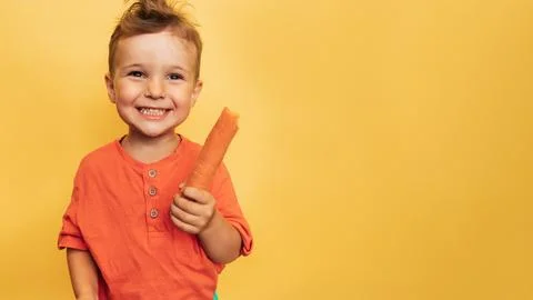 Studio shot of a smiling boy holding a fresh carrot on a yellow background. The Stock Photos