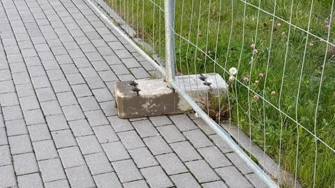 Sturdy grey concrete block secures metal fence on a varied texture pavement.. Stock Photos