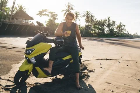 Stylish barefooted guy using smartphone sitting on motorbike in tropical resort Stock Photos