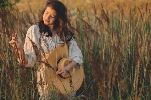 Stylish boho woman with straw hat posing among wild grasses in evening. Sum.. Stock Photos
