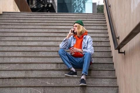 Stylish guy with coffee cup in hand sits on steps talks on smartphone in city Stock Photos