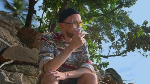 Stylish guy in yellow glasses sitting on rocky beach smoking joint or cigarette Stock Footage 236839655