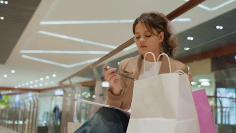 Stylish influencer sorting goods on bench during mall visit, young fashion lover Stock Footage 326100614