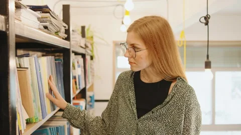 In a stylish library, a focused, red-haired female student is immersed in her Stock Footage 285091680