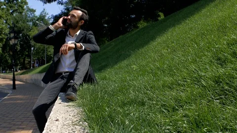 Stylish man with a beard sitting on a concrete wall and talking on the phone Stock Footage 113965693