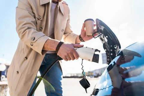 Stylish man inserts plug of the charger into the socket of electric car Stock Photos