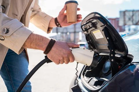 Stylish man inserts plug of the charger into the socket of electric car Stock Photos