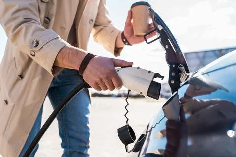 Stylish man inserts plug of the charger into the socket of electric car Stock Photos