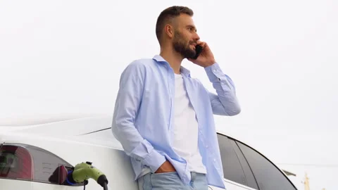 Stylish man phoning while charging car at electric vehicle charging station Stock Footage 251763831