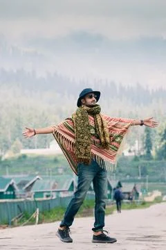 A stylish man wearing a patterned poncho, a scarf, and a hat stands with his  스톡 사진