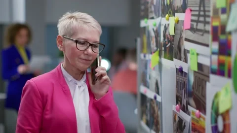 Stylish mature editor talking on smartphone standing near mood board in creative Stock Footage 189230730