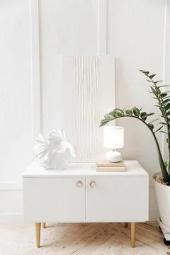 Stylish white bedside table with decor, lamp and white abstract gypsum painting Stock Photos
