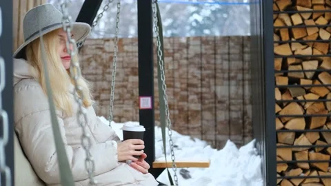 Stylish woman drinking coffee while sitting on a swing in winter Stock Footage 163308831