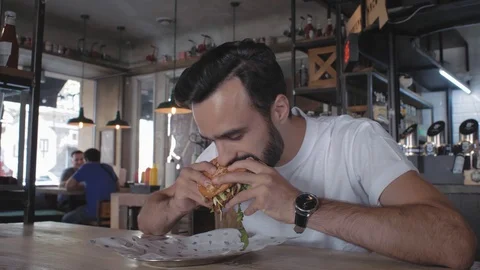 Stylish young guy with a beard bites a burger sitting at a table in a cafe. 4k Stock Footage 115634216