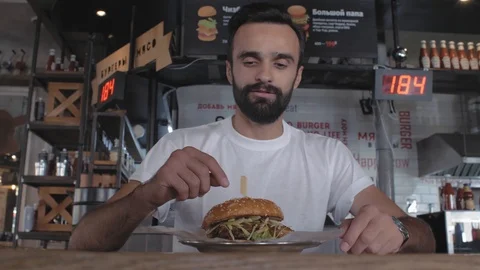 Stylish young guy with a beard sits in a cafe with a plate with a burger. 4k Stock Footage 115634134