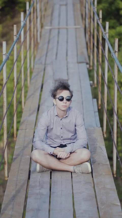 A stylish young guy in round sunglasses and a light checkered shirt sits cross Stock Footage 345528462