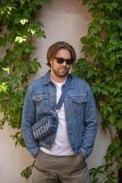A stylish young man in a denim jacket and sunglasses poses confidently agains Фото