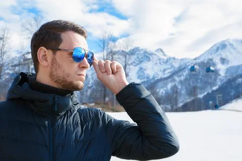 Stylish young man in down jacket holding sunglasses in front of ski lifts and Фото