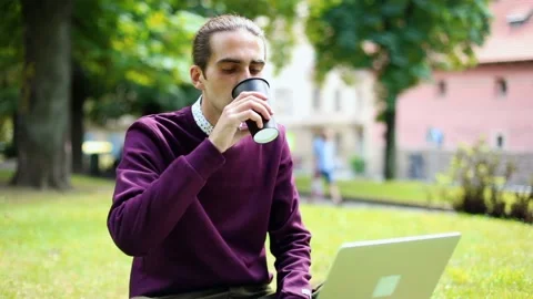 Stylish young man using laptop drinking coffee networking online in city park. U 스톡 동영상 145756259