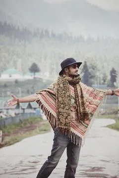 A stylish young man wearing a patterned poncho, scarf, and hat standing with  스톡 사진